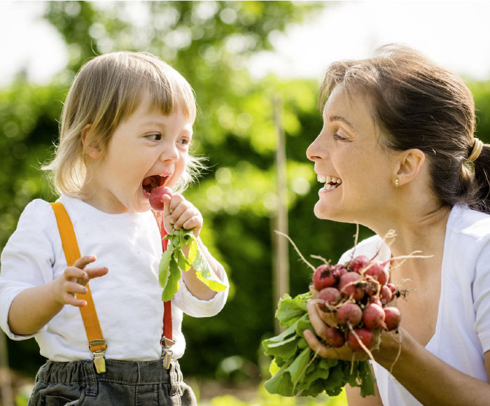 A young child bites a radish with its mother looking on smiling