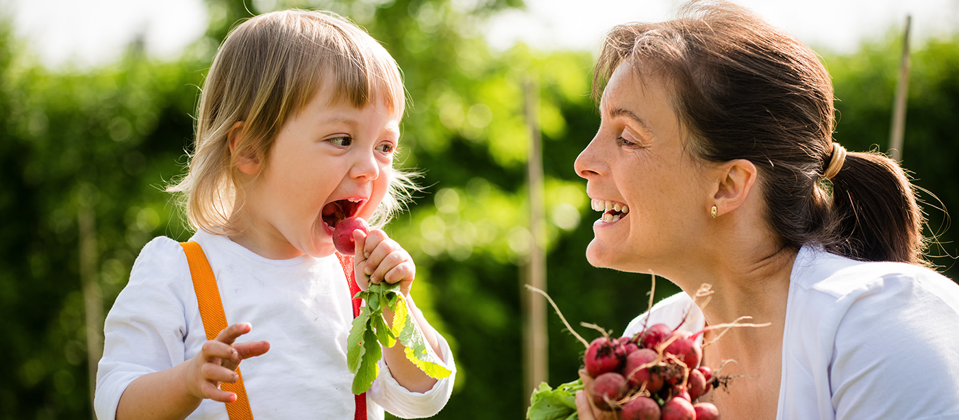 a little girl about to bite a radish with her mother smiling watching her